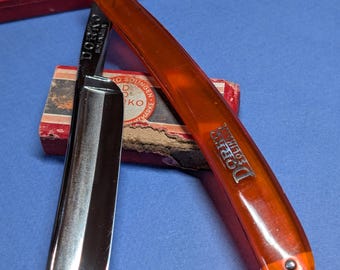 May include: A vintage straight razor with an orange handle and a silver blade, lying on a blue surface. The razor is marked "DORCO SOLINGEN". A red and white box is in the background.