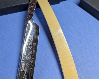 May include: A vintage straight razor with a cream-coloured handle and a silver blade. The blade has etched text including "Svenskt Diamant Stål Allmänco Spezial" and "Tio Års Garanti". The razor is displayed on a blue surface with a black box.