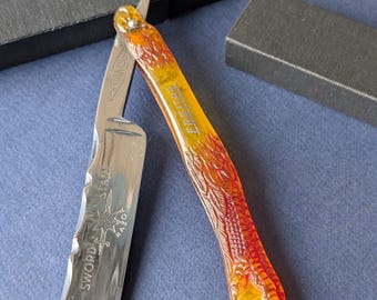 May include: A straight razor with a polished steel blade and an orange and red handle. The handle is etched with a decorative pattern. The blade is marked with the words "SWORD STAR RAZOR". The razor is open, and the blade is visible.