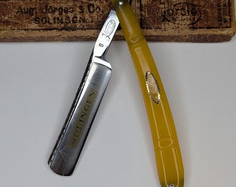 Puede incluir: Una navaja de afeitar abierta con mango amarillo y hoja plateada. La hoja está grabada con la palabra "SOLINGEN". La navaja se encuentra frente a una caja marrón con el texto "Aug. Jörges & Co. SOLINGEN".