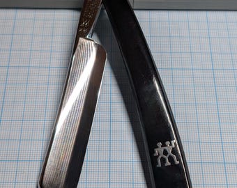 May include: A black straight razor with a silver blade is open, revealing the sharp edge. The handle features a silver emblem of two figures. The razor is positioned on a blue grid surface, with a gray case in the background.