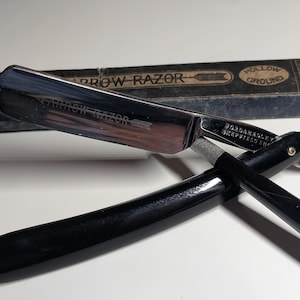 May include: A vintage straight razor with a black handle and a silver blade, labelled "ARROW RAZOR." The razor is open, displaying the sharp edge. A rectangular box with text is in the background.