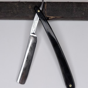 以下が含まれることがあります： 黒いハンドルと銀色の刃が付いた、開いた状態のヴィンテージストレートカミソリ。ハンドルには金色のリベットが付いています。カミソリは、テキストの入ったぼやけた茶色の革ケースの前に斜めに配置されています。