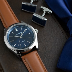 May include: A silver watch with a blue face and brown leather band. The watch face has the words "Langdon" and "Jubilee" printed on it. The watch is lying on a brown surface with a blue bow tie and silver cufflinks.
