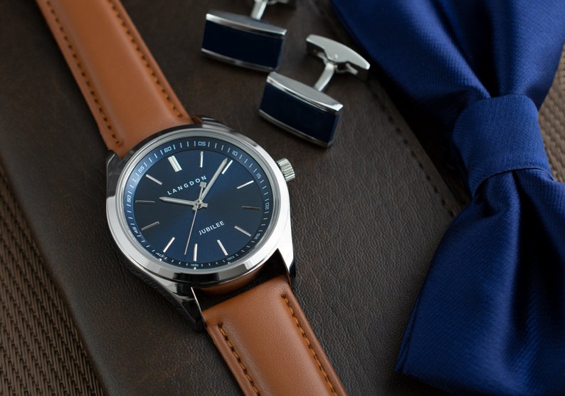 May include: A silver watch with a blue face and brown leather band. The watch face has the words "Langdon" and "Jubilee" printed on it. The watch is lying on a brown surface with a blue bow tie and silver cufflinks.