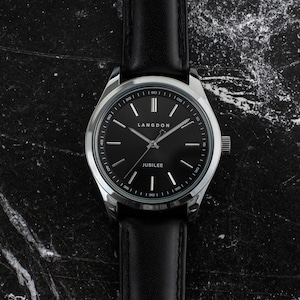 May include: A silver watch with a black leather band and a black face. The watch face has white markings and the words "LANGDON" and "JUBILEE" printed on it.