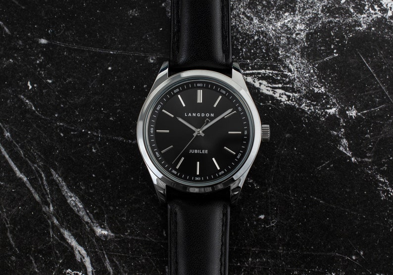 May include: A silver watch with a black leather band and a black face. The watch face has white markings and the words "LANGDON" and "JUBILEE" printed on it.