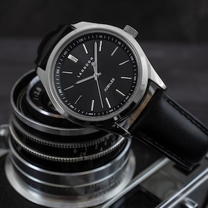 May include: A silver watch with a black leather band and a black face. The watch face has white markings and the words "Langdon" and "Jubilee" printed on it.