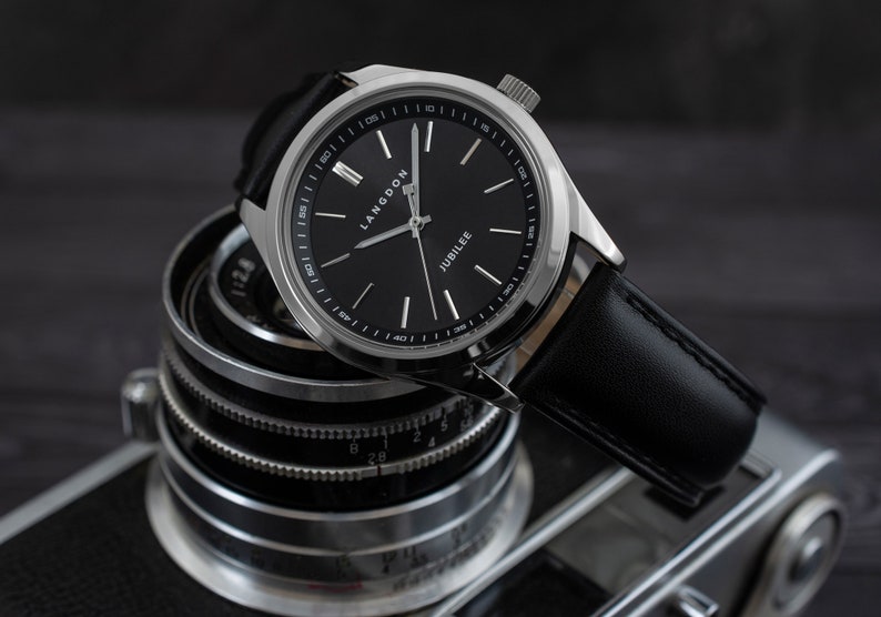 May include: A silver watch with a black leather band and a black face. The watch face has white markings and the words "Langdon" and "Jubilee" printed on it.