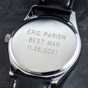 May include: A silver watch with a black leather band. The back of the watch is engraved with the text "Eric Parish Best Man 11.26.2021".