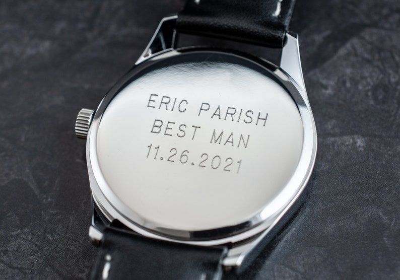 May include: A silver watch with a black leather band. The back of the watch is engraved with the text "Eric Parish Best Man 11.26.2021".