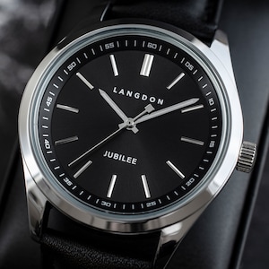 May include: A silver watch with a black face and black leather band. The watch face has white markings and the words "LANGDON" and "JUBILEE" printed on it.