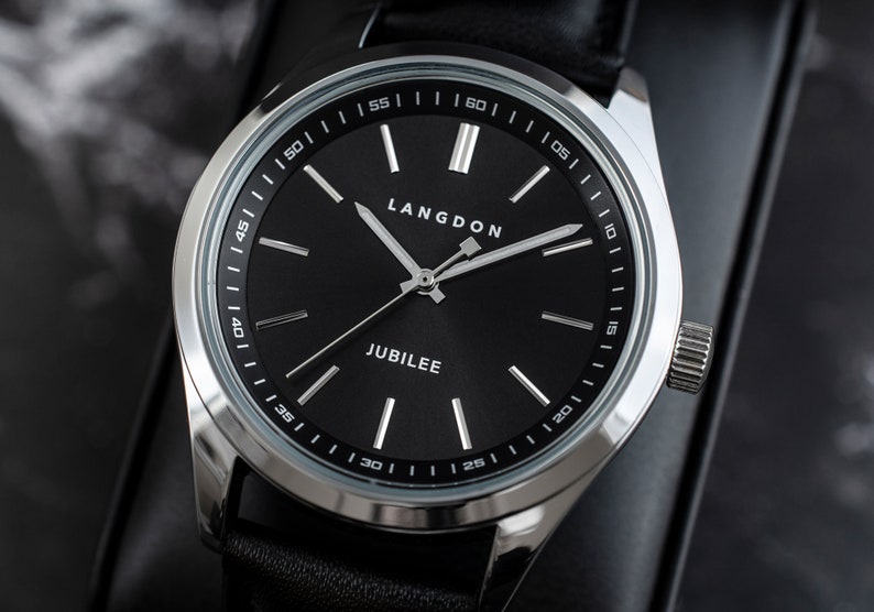 May include: A silver watch with a black face and black leather band. The watch face has white markings and the words "LANGDON" and "JUBILEE" printed on it.