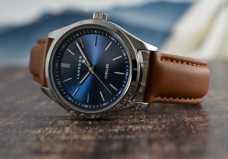 May include: A silver watch with a blue face and brown leather band. The watch face has the words "Langdon Jubilee" printed on it.