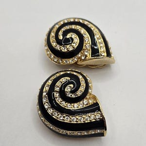 Peut inclure: Paire de boucles d'oreilles vintage à clip en forme de coquillage. Les boucles d'oreilles présentent une base en émail noir avec un motif en spirale doré, incrusté de strass transparents. Elles ont un fermoir à clip doré.
