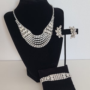 Könnte beinhalten: Ein dreiteiliges Schmuckset aus silberfarbenem Strass, bestehend aus einer Halskette, einem Armband und Ohrringen. Die Halskette ist mehrlagig mit einer abgestuften Länge aus Strasssteinen. Das Armband ist einreihig mit Strasssteinen. Die Ohrringe sind klein und haben einen Strassstein-Cluster.