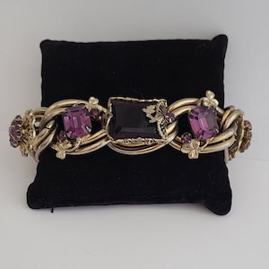 May include: A gold-tone bracelet with a chain link design and purple glass stones. The bracelet has a floral motif and is accented with smaller stones.