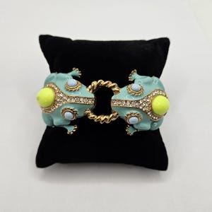 Könnte beinhalten: Ein Klapparmband mit zwei hellblauen Froschfiguren, die sich gegenüberstehen und durch ein gedrehtes, goldfarbenes Metallglied verbunden sind. Jeder Frosch ist mit leuchtend gelben Cabochon-Augen, hellblauen Rund-Einlagen und klaren Strasssteinen verziert.