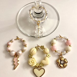 May include: Set of four wine glass charms. Each charm features a different design: a lightning bolt, a heart, a flower, and a crystal bead. The charms are made of gold-tone metal and beads.
