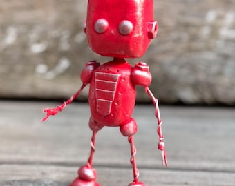 May include: A small, hand-crafted robot figurine featuring a bright red, rounded head with two metallic silver eyes. The torso is also red with a silver-accented chest plate, while the thin arms and legs are constructed from twisted red wire.