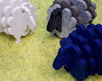 May include: Three felt sheep coasters in white, grey, and blue. The sheep are stacked on top of each other and have a 3D design.