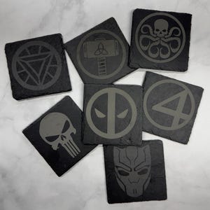 May include: A collection of square, dark gray slate coasters. Each coaster features a different superhero or villain symbol, including Iron Man's arc reactor, Thor's hammer, and the Hydra logo. The designs are etched in a lighter gray.