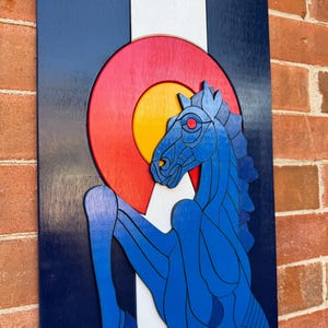 Puede incluir: Arte mural de madera con el dise&ntilde;o de la bandera del estado de Colorado. La obra de arte incluye un caballo azul encabritado, un emblema circular rojo y amarillo, y franjas verticales azules, blancas y azules. La pieza est&aacute; montada en una pared de ladrillo.
