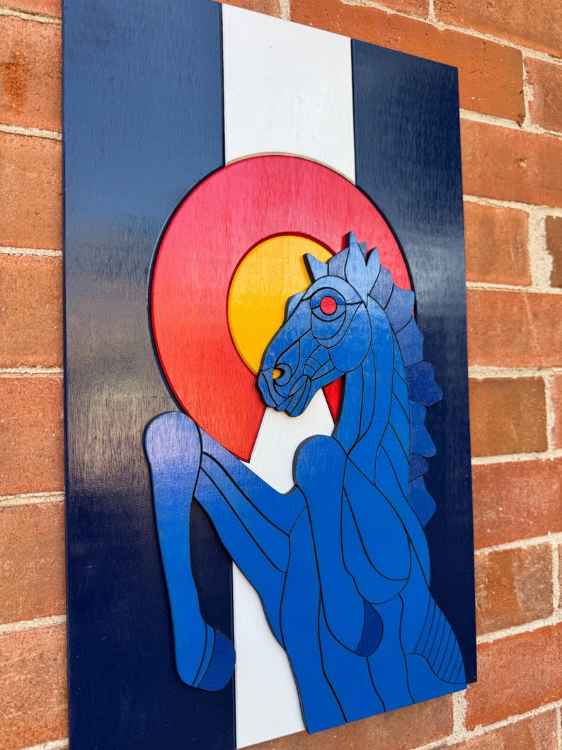 Puede incluir: Arte mural de madera con el dise&ntilde;o de la bandera del estado de Colorado. La obra de arte incluye un caballo azul encabritado, un emblema circular rojo y amarillo, y franjas verticales azules, blancas y azules. La pieza est&aacute; montada en una pared de ladrillo.