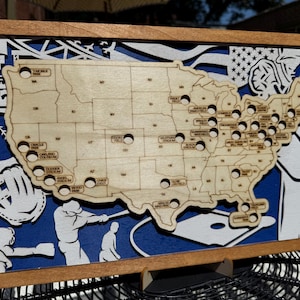May include: Wooden baseball-themed wall art featuring a laser-cut map of the United States with stadium locations. The artwork includes baseball players, a glove, and an American flag design, all set against a blue background within a wooden frame.