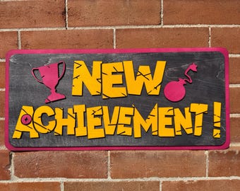 May include: A rectangular sign with a dark gray background and a pink border. The sign features the words "NEW ACHIEVEMENT!" in yellow, along with pink illustrations of a trophy and a bomb.