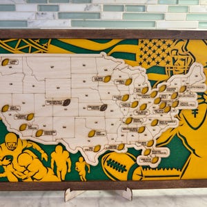 May include: Wooden map of the United States with football-shaped markers indicating stadium locations. The map is set against a green and yellow background with football-themed graphics and a brown frame. The map is approximately 24 inches wide and 14 inches tall.