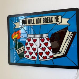 May include: A framed art piece with a blue background and black border. The artwork features a pair of white shorts with red hearts, a bottle with a flame, a book, and the text "YOU WILL NOT BREAK ME" on a banner.