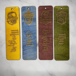 May include: Four wooden bookmarks with engraved designs and names. Each bookmark features a different color: yellow, blue, red, and green. The bookmarks have a hole at the top for a string. The designs include the Hogwarts houses: Hufflepuff, Ravenclaw, Gryffindor, and Slytherin.