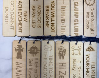 May include: A collection of wooden bookmarks with various engraved designs and phrases. Each bookmark has a dark blue tassel. The phrases include "God Damnit Donut!", "Achievement", and "You Will Not Break Me".