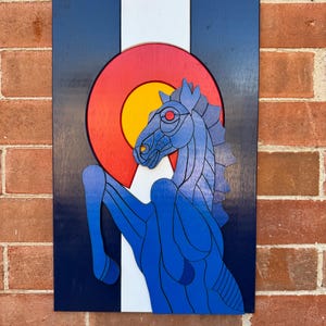 Puede incluir: Arte mural de madera con la bandera del estado de Colorado y un caballo azul. La obra de arte tiene un fondo azul marino con una franja blanca y un c&iacute;rculo rojo y amarillo. El caballo es azul con ojos rojos.