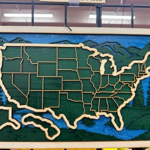 May include: Wooden wall art depicting the United States map. The map is layered with a green base, tan state outlines, and blue accents for water and mountains. The frame is a light tan color, and the overall design is a 3D effect.