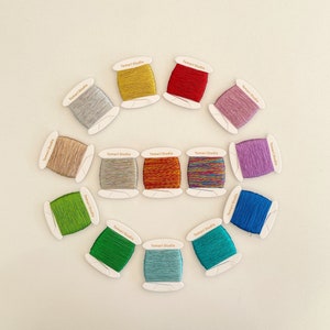 Op de afbeelding: Een verzameling van 15 spoelen borduurgaren in verschillende kleuren, waaronder goud, rood, roze, paars, blauw, groen en bruin. Elke spoel is gelabeld met "Temari Studio".