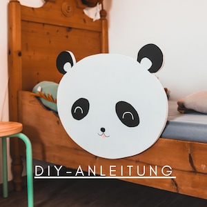 Puede incluir: Una cara de panda blanca y redonda con ojos y orejas negras y una boca rosa. El panda está colocado frente a una estructura de cama de madera. El texto "DIY-ANLEITUNG" está escrito en blanco en la parte inferior de la imagen.