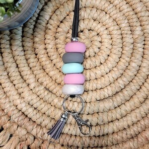 Puede incluir: Un cordón negro con un llavero plateado y un borla gris. El cordón tiene cuatro cuentas de colores: rosa, gris, azul claro y rosa.