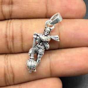 May include: A silver-toned metal pendant featuring a detailed figure of a deity holding a large mace. The figure is depicted in a dynamic pose with a flowing garment and a prominent bail at the top for attachment to a chain or cord.