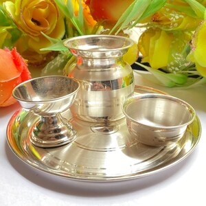 May include: A polished silver-toned metal set consisting of a small round tray, a central pot with a flared rim, a pedestal-based bowl, and a simple small bowl. The items feature a smooth, reflective surface with subtle horizontal line detailing.