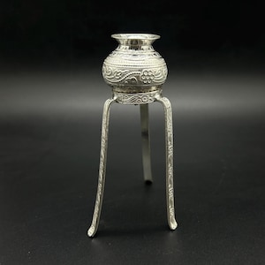 May include: A silver decorative stand with a round top and three legs. The stand is decorated with an intricate floral pattern.