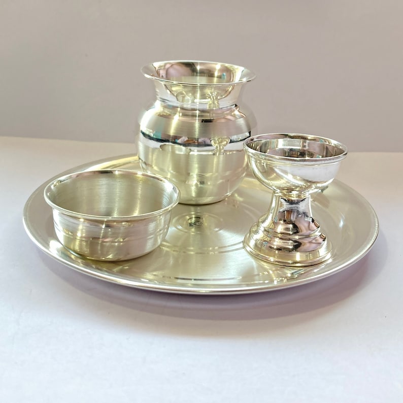 May include: A set of three polished silver-coloured metal items consisting of a round tray, a small bowl, a wide-mouthed pot, and a pedestal-based cup. Each piece features a smooth, reflective surface with a metallic finish and simple, clean lines.