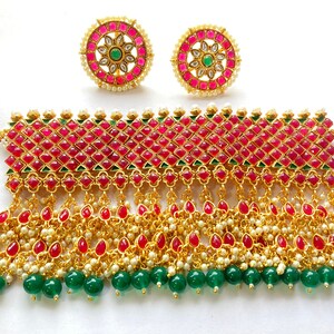 May include: A gold choker necklace with a red and green stone design. The necklace has a thick band with a row of red stones and a row of green stones. The band is attached to a long chain with red and green stones. The necklace is paired with matching earrings.