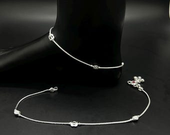 May include: A pair of delicate silver anklets. One anklet is around the ankle, and the other is lying on a black surface. Each anklet features small, decorative elements and a chain-like design. The anklets are displayed on a black mannequin foot.