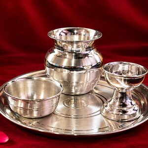 May include: A polished silver-toned metal set consisting of a round tray, a small bowl, a rounded pot with a flared rim, and a pedestal-style cup. The items feature smooth, reflective surfaces with subtle horizontal etched lines for decoration.