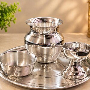 May include: A set of polished silver-coloured metal items including a round tray, a small bowl, a wide-mouthed pot with a curved neck, and a pedestal-based cup. Each piece features a smooth, reflective surface and simple, clean lines.