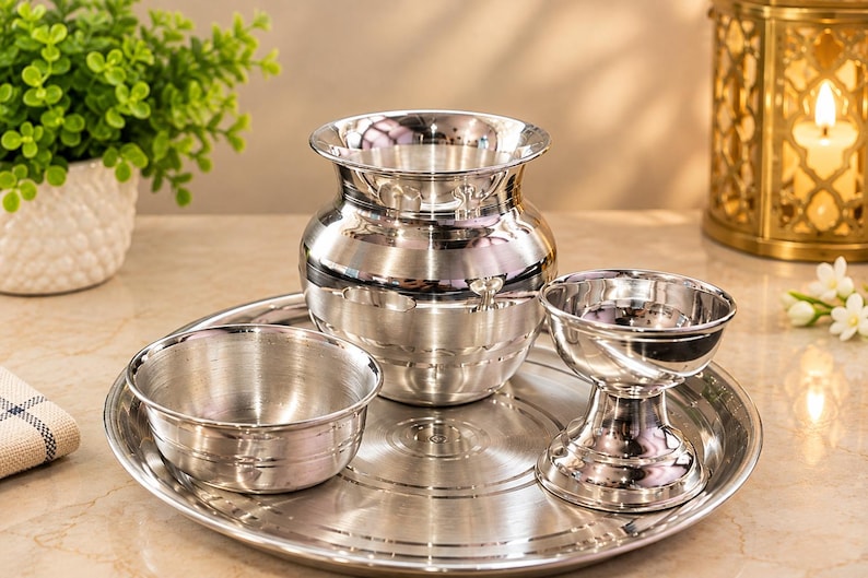 May include: A set of polished silver-coloured metal items including a round tray, a small bowl, a wide-mouthed pot with a curved neck, and a pedestal-based cup. Each piece features a smooth, reflective surface and simple, clean lines.