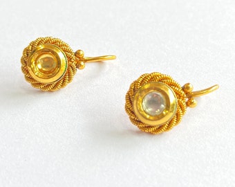 Op de afbeelding: Een paar gouden oorbellen met een ronde, witte steen die in een gouden zetting is gezet. De oorbellen hebben een gedraaid draadontwerp en een kleine haaksluiting.
