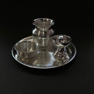 May include: A polished silver-toned metal set consisting of a circular tray, a small bowl, a rounded pot with a flared rim, and a pedestal-based cup. Each piece features a smooth, reflective surface and a simple, clean design for functional use.